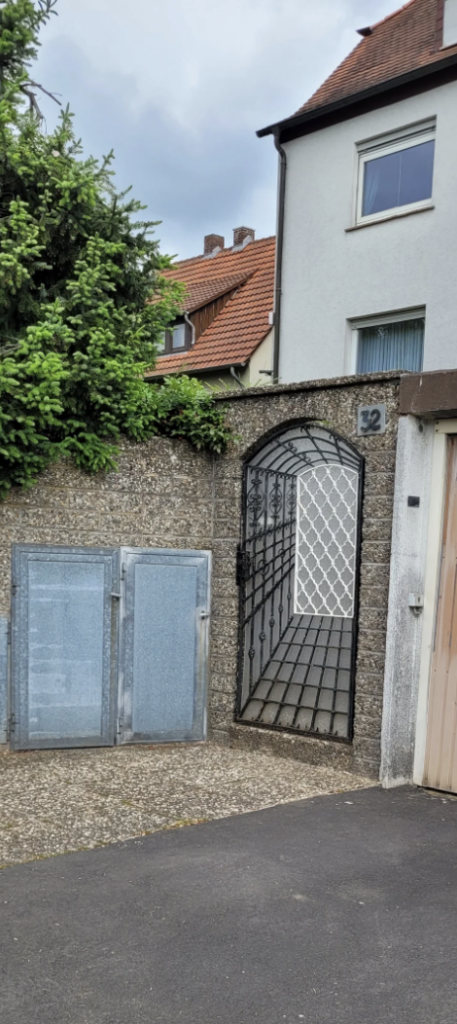 Image of a residential entrance showing a wrought iron gate situated in a stone wall, alongside a white building with a red tiled roof. There are green shrubs above the wall, and two covered electrical or utility boxes visible to the left of the gate. House number 32 is visible.