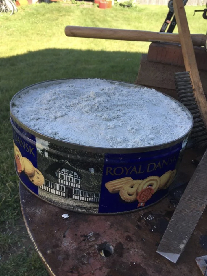 Safety fails that show a Royal Dansk cookie tin filled with white ash is placed on a wooden workbench outside. There are some tools around the bench, including a broom and a yardstick. A lawn and some outdoor items can be seen in the background.