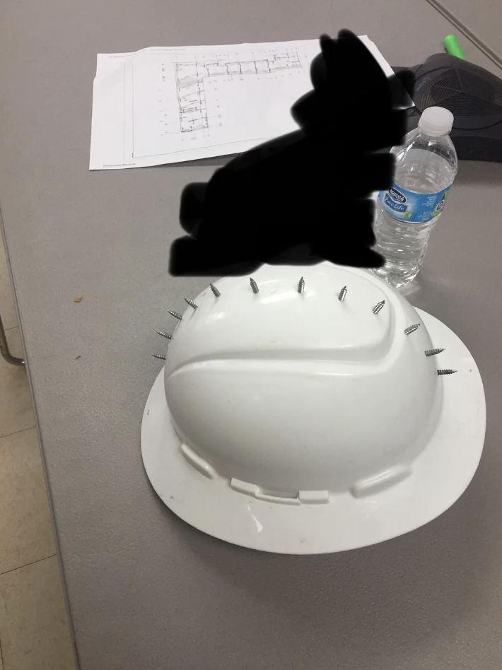 A white construction hard hat with several screws protruding from it is placed on a gray table. A water bottle and some papers with diagrams are also on the table. Parts of the image are blacked out.