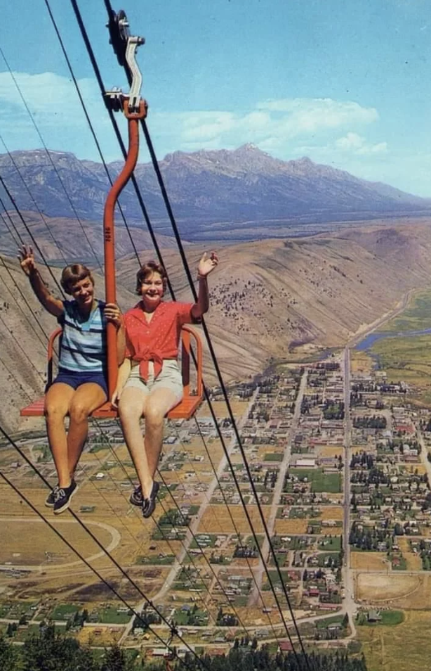 Two women sit on a red chairlift, smiling and waving, high above a town nestled in a valley. The town is surrounded by expansive, rocky hills and majestic mountains under a clear, blue sky. The scene captures a sense of adventure and scenic beauty.