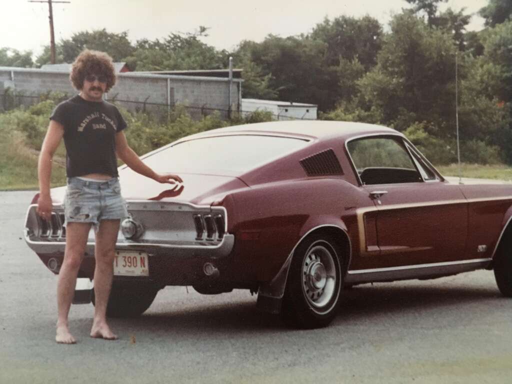 A man with curly hair, sunglasses, and a mustache stands barefoot on a paved area. He is wearing a black t-shirt and denim shorts. He is touching the trunk of a maroon vintage car, likely a Ford Mustang, with trees and a fence in the background.