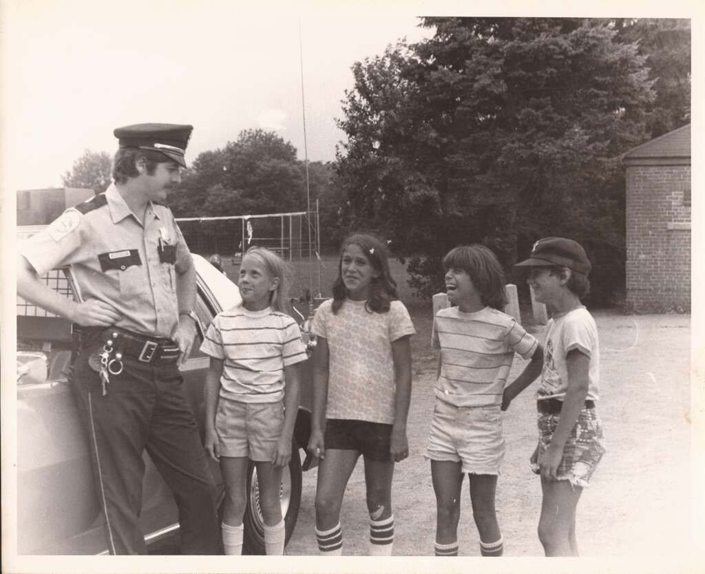 A police officer stands next to a patrol car, engaging in conversation with four children. The children, dressed in casual summer clothing with striped socks, appear engaged and happy. In the background, there are trees and a brick building. The image is in black and white.