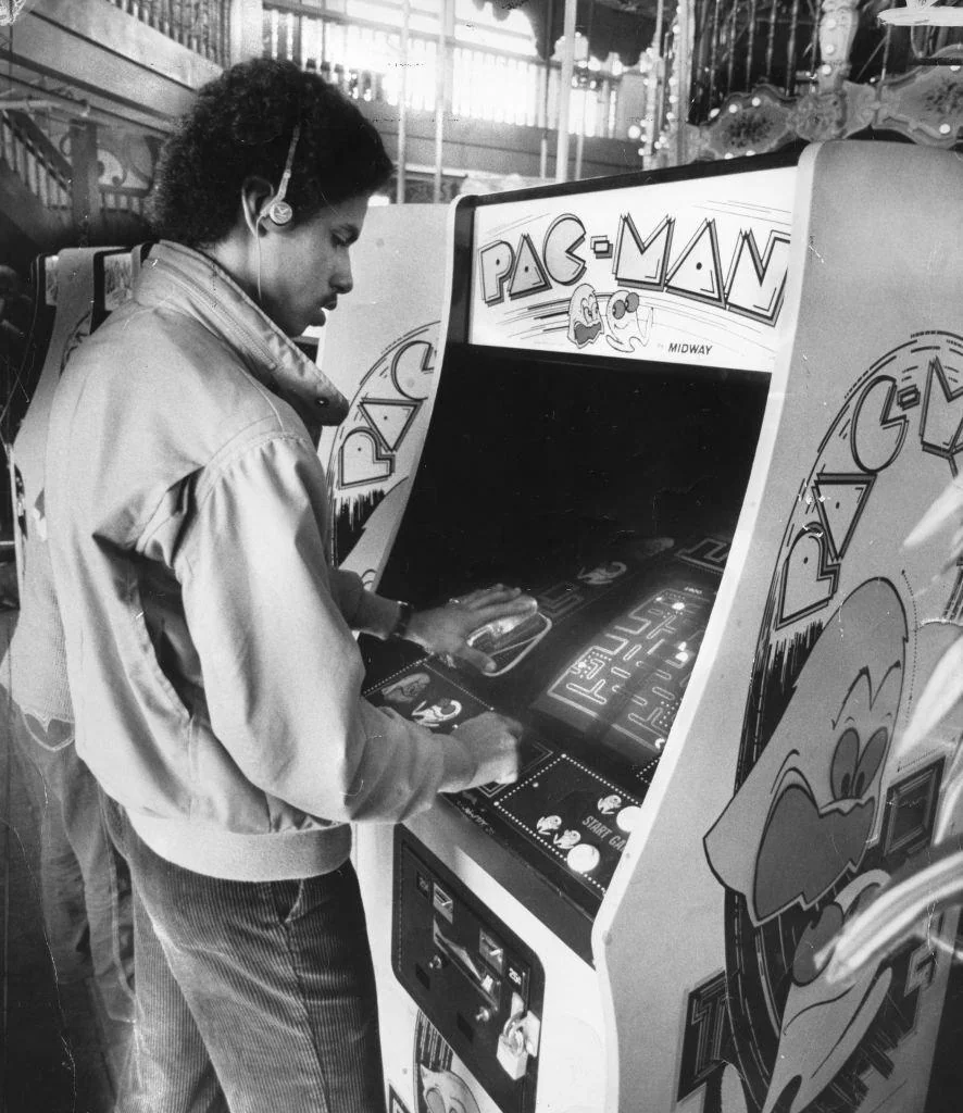 A person is playing a Pac-Man arcade game in a vintage arcade setting. The person is focused on the screen, using controls with both hands. The arcade machine has colorful artwork, including the Pac-Man logo and character graphics. Another person is nearby.