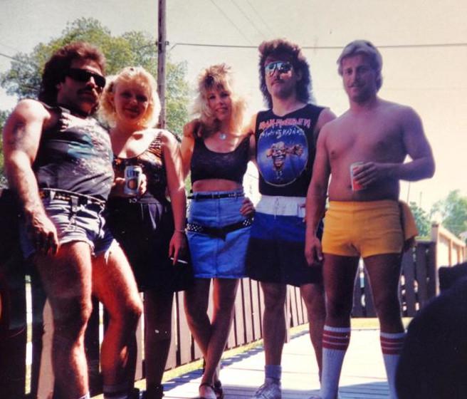 A group of five people stand on an outdoor wooden deck. They are dressed in 80s attire, including shorts, tank tops, and sunglasses. Two men on the left have mustaches and the man on the far right is shirtless. They all appear to be posing and enjoying themselves.