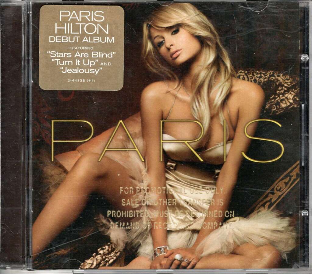 The image shows the cover of Paris Hilton's debut album. She is posing in a glamorous setting wearing a beige dress. The cover features text that reads: "PARIS" and "Paris Hilton Debut Album featuring 'Stars Are Blind,' 'Turn It Up,' and 'Jealousy.'