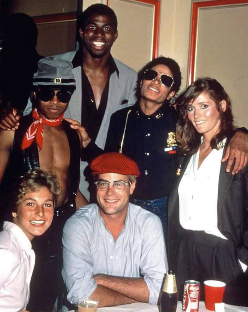 A group of people, including two men standing in the back wearing sunglasses and hats, and three others seated at a table with beverages, smile for the camera. The diverse group is casually dressed and appears to be enjoying a social gathering indoors.