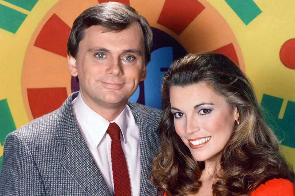 A man and woman are standing closely, smiling at the camera. The man is wearing a grey suit jacket, white shirt, and red tie. The woman has long brown hair and is wearing a red top. The background is colorful with concentric patterns.