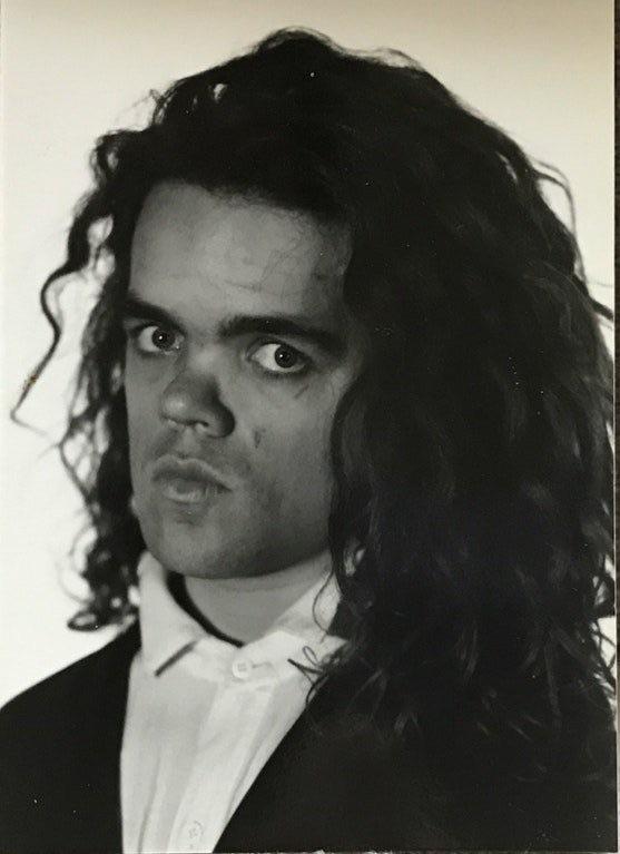 Black and white portrait of a person with long, curly hair, staring directly at the camera. They are wearing a white shirt and a dark jacket. The expression on their face is serious.