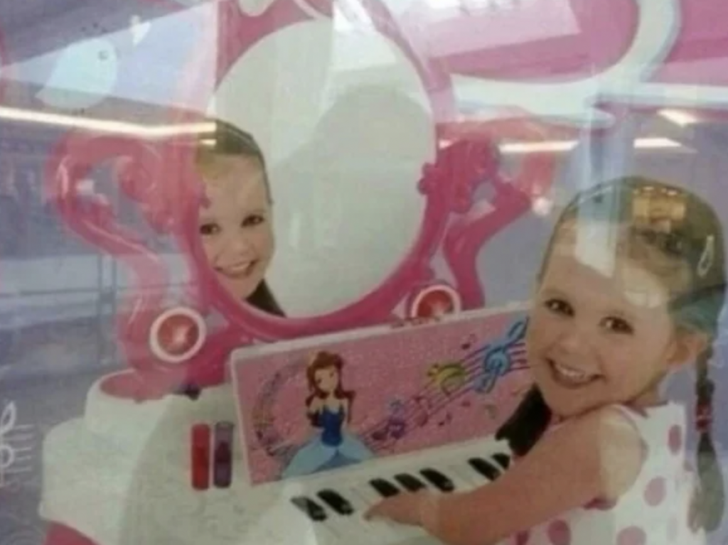 A young girl with brown hair and a pink dress smiles while playing on a pink toy piano with a music-themed design and a figure of a princess. Her reflection is visible in an attached white and pink vanity mirror.