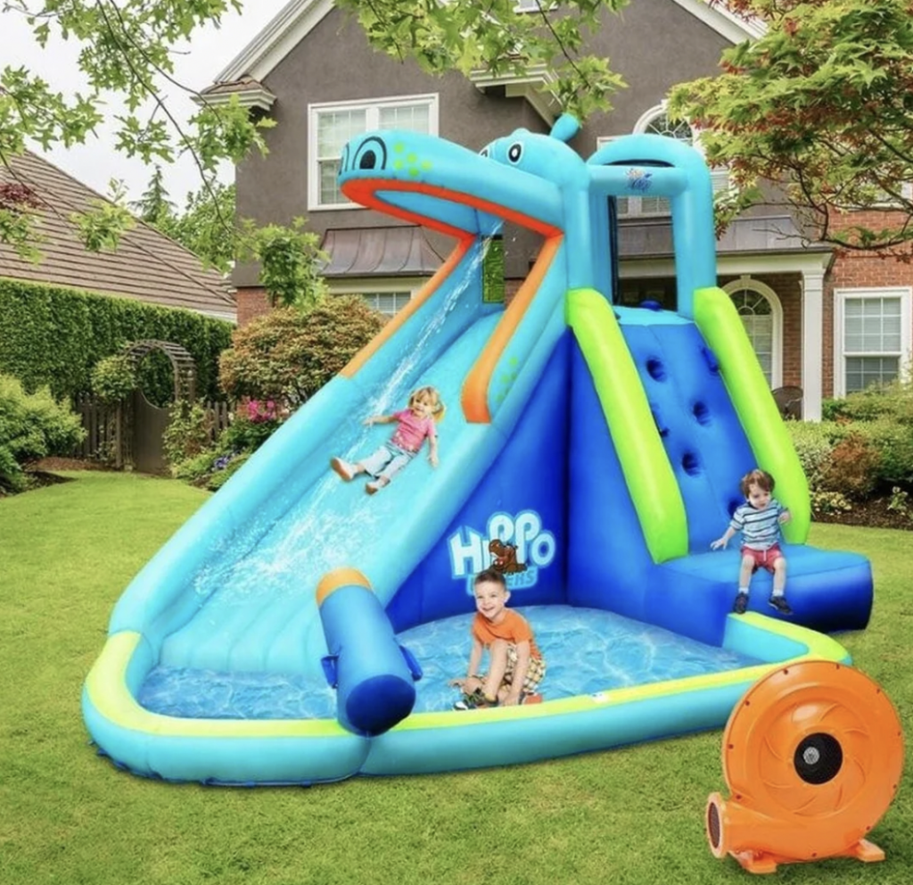 Three children play on an inflatable water slide in a backyard. The slide has a climbing wall and a pool at the bottom. The children look happy as they splash around. The house and garden are visible in the background, and an air blower is in the foreground.