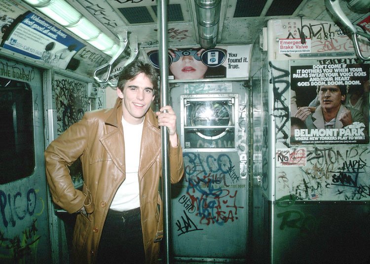 A person wearing a brown leather jacket stands inside a graffiti-covered subway car, holding onto a metal pole. The walls and doors are heavily marked with colorful scribbles and tags. The space around him is adorned with vintage posters and advertisements.