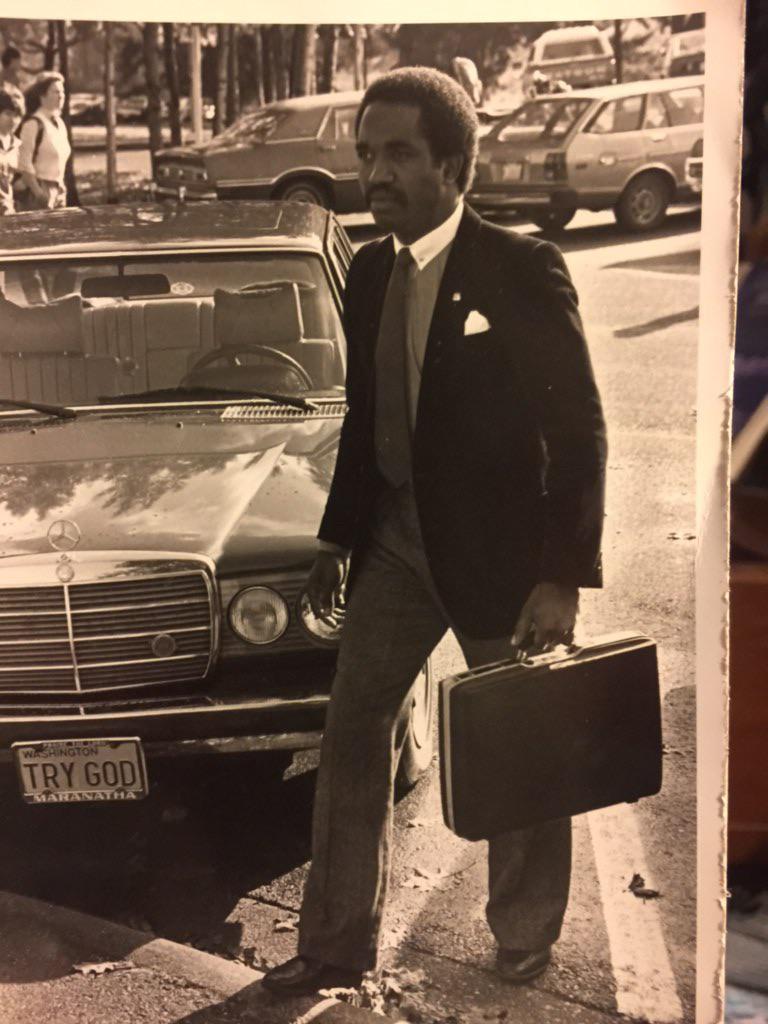 A man dressed in a suit and tie walks through a parking lot while holding a briefcase. He is passing a Mercedes-Benz car with a license plate that reads "TRY GOD." In the background, there are more parked cars and trees. The image is in black and white.