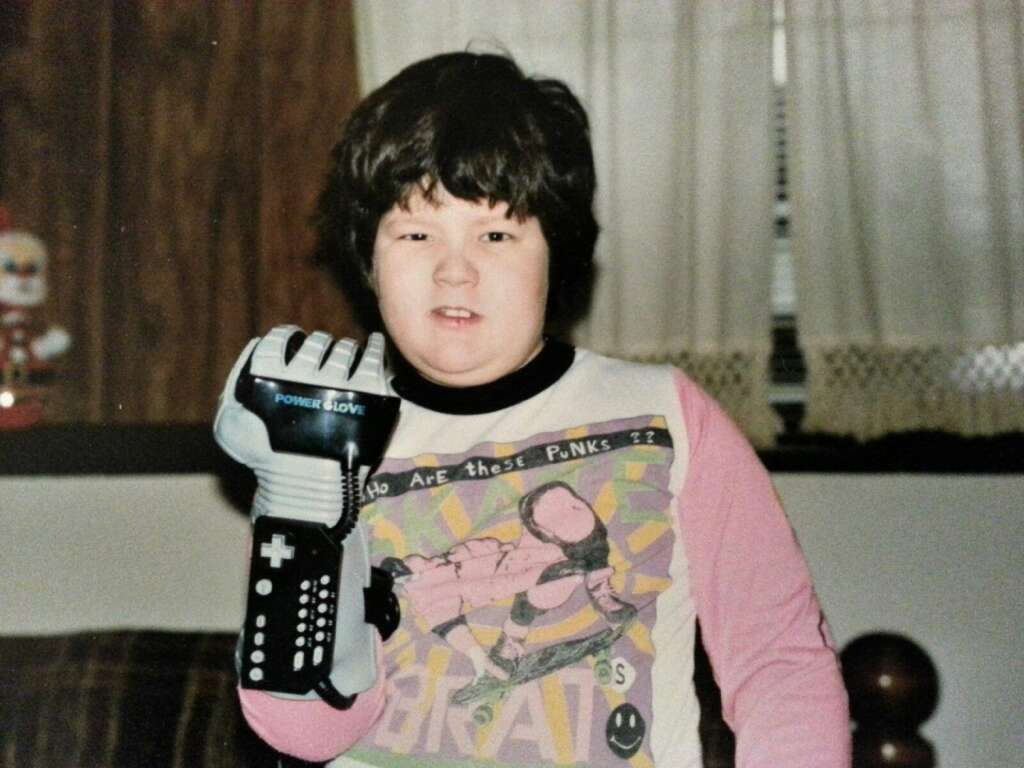 A young child stands indoors, wearing a shirt with a cartoon on it and holding up a gloved hand. The glove is a Nintendo Power Glove, a video game controller from the 1980s. The child has dark hair and is smiling slightly, appearing excited or proud.