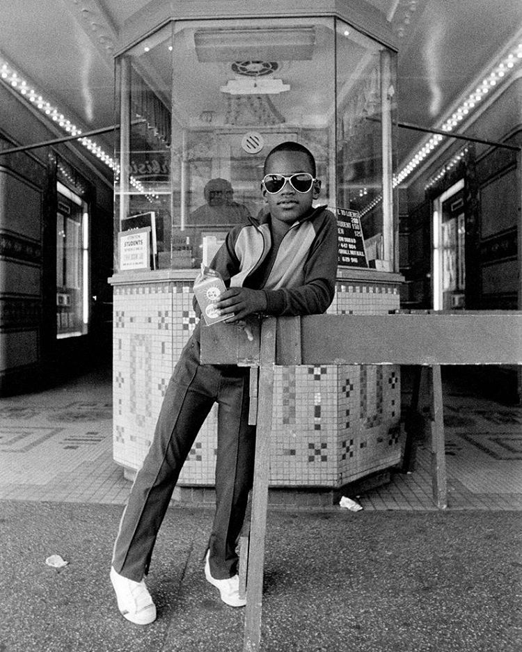 A young boy wearing sunglasses and a tracksuit leans against a barricade in front of a building entrance. He holds a soda cup and bag of popcorn. The setting appears to be a vintage movie theater with a tiled ticket booth and a well-lit interior.