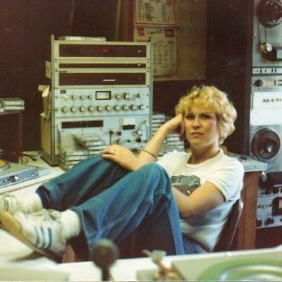 A person with short blonde hair is sitting in a relaxed manner at a desk, with their legs up on a chair, in a radio station or recording studio. They are wearing a white shirt, blue jeans, and white sneakers. Various audio equipment and a calendar are visible in the background.