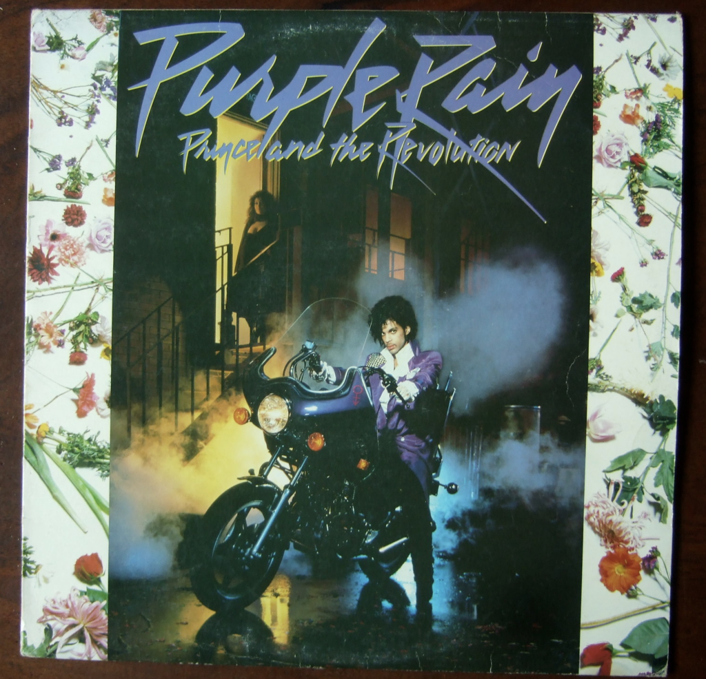 Album cover for "Purple Rain" by Prince and The Revolution. The image features Prince, dressed in purple, sitting on a motorcycle with steam around him. The background shows a dark street scene with buildings and the title "Purple Rain" in stylized text at the top.