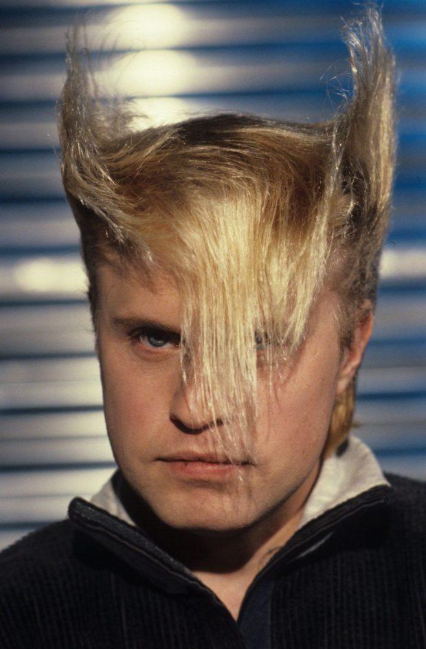 A person with blonde hair styled into pointy, upward spikes on either side of their head and long bangs obscuring part of their face, stares intently at the camera. The background consists of horizontal blinds, adding texture to the image.