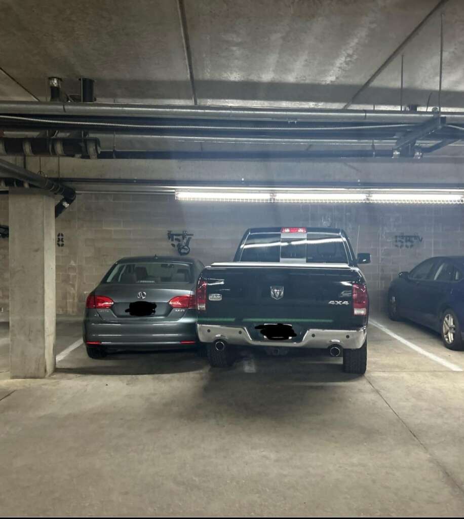 A dimly lit underground parking lot with two vehicles parked closely next to each other. On the left is a grey car and on the right is a green pickup truck, both with partially unclear license plates. Another vehicle is faintly visible to the right.