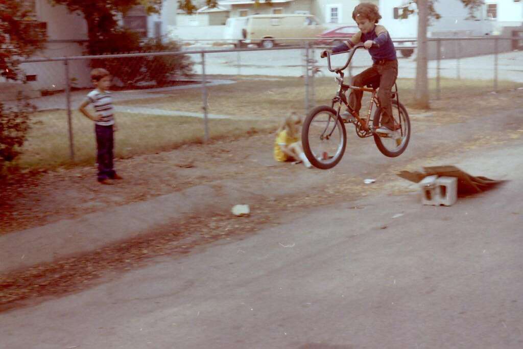 A young child is airborne on a bicycle, jumping over a makeshift ramp made of a board and a cinder block on a suburban street. A boy stands to the left watching, while a girl in a yellow dress sits on the grass nearby. A fence and houses are visible in the background.