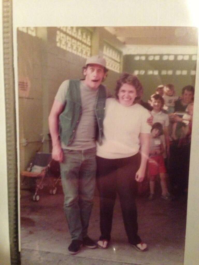 Two people standing together inside a building with patterned walls. The person on the left is wearing a cap, a sleeveless vest, and looks surprised with their mouth open. The person on the right, smiling, is wearing a white shirt. A crowd is in the background.