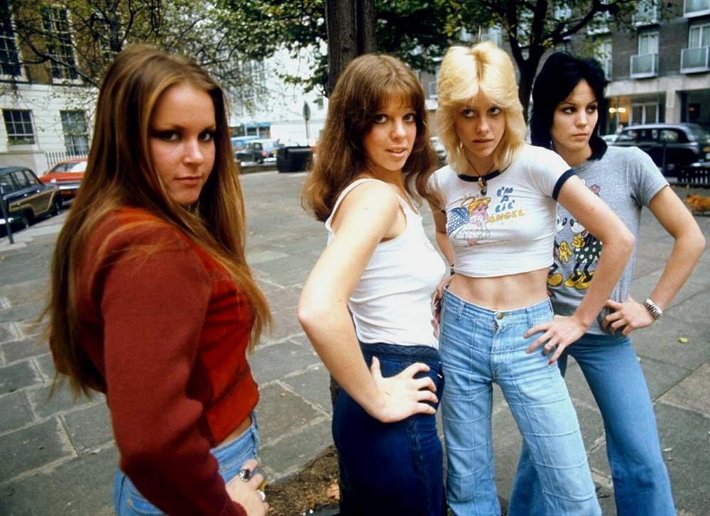 Four women pose confidently on a sidewalk in an urban setting with trees and buildings in the background. Three have long hair, and one has short, black hair. They wear casual 1970s-era clothing, including flared jeans and graphic t-shirts.