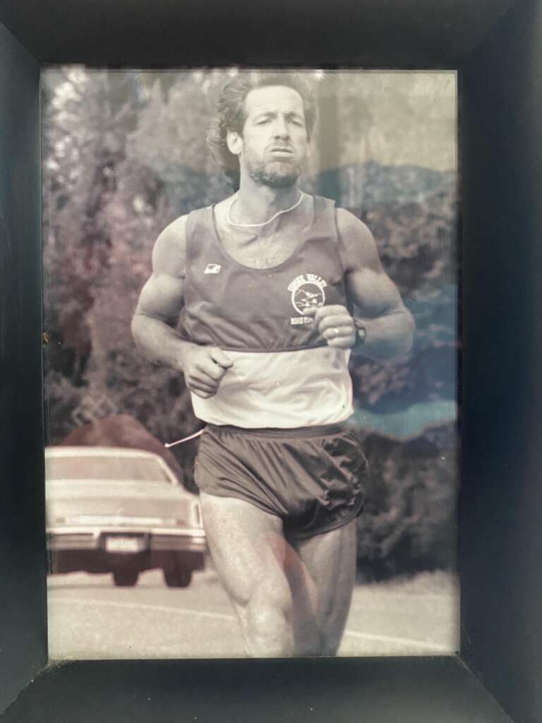 Black and white photo of a male runner in athletic gear, jogging outdoors. He is wearing a tank top and shorts, and appears focused and determined. A vintage car is visible in the background, along with trees and outdoor foliage.