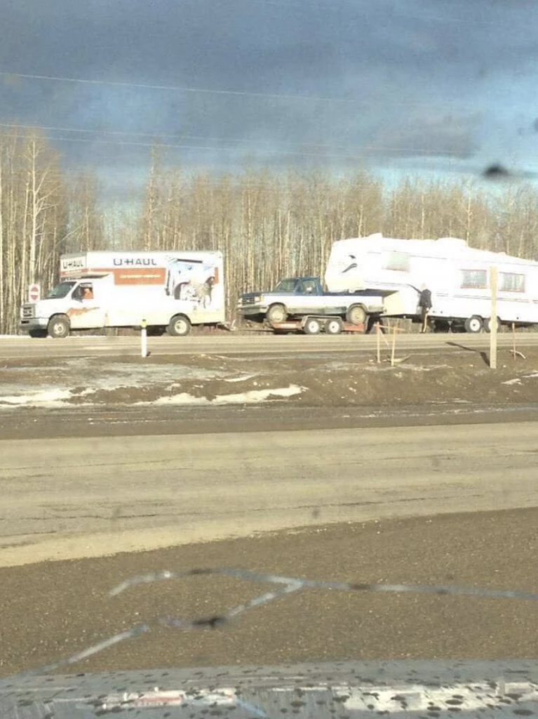A U-Haul truck is pulling a trailer carrying a car and an RV on a road surrounded by bare trees and patches of snow. The scene is captured from a vehicle moving alongside, with part of the interior visible in the foreground.