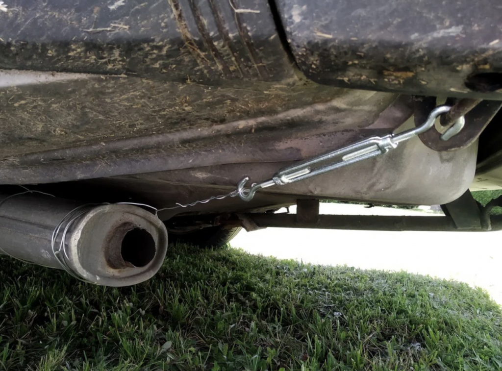 A car's exhaust pipe is secured with a wire and what appears to be a turnbuckle, attached to the undercarriage of the vehicle above grassy ground. The components show signs of wear and dirt, suggesting a makeshift repair.