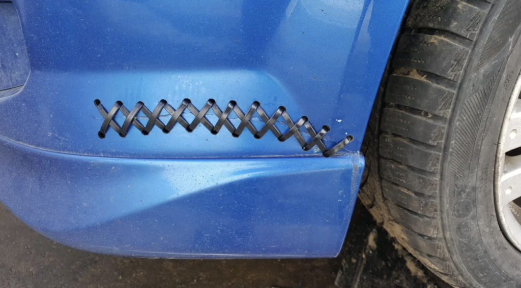 A close-up view of a blue car's bumper with a series of zip ties securing a long vertical crack. The crack runs from the bottom of the bumper up to just below the hood, and the black zip ties form a zigzag pattern to hold the damaged pieces together.
