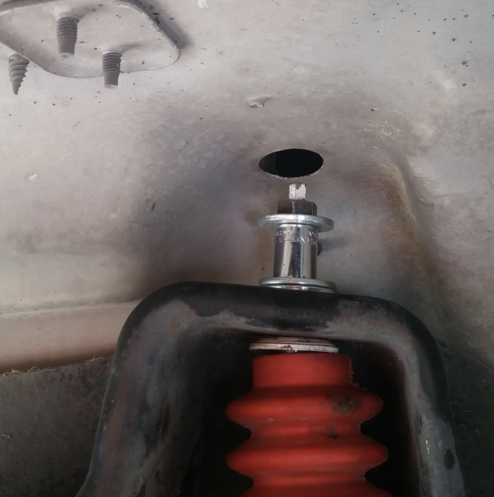 Close-up of a car's suspension system, focusing on a red coil spring seated in a black suspension mount. Above the mount, a metal bolt is partially visible through a hole in the vehicle's chassis. The surrounding area appears to be part of the wheel well.