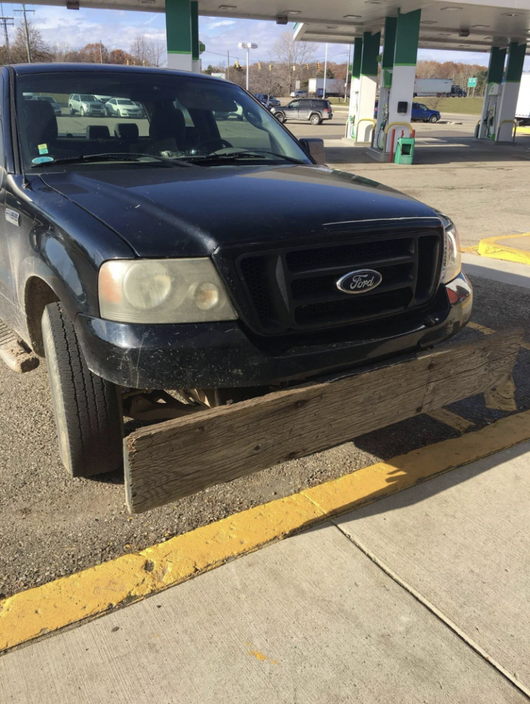 A black Ford truck is parked at a gas station with a large wooden plank attached to its front bumper. The day appears sunny with a clear sky, and the ground is dry. The gas station has some empty parking spaces and visible gas pumps in the background.
