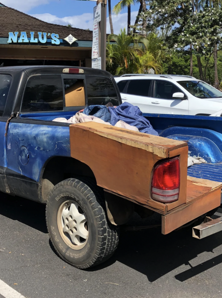 A blue and black pickup truck with a wooden extension on its bed is parked in a lot. The truck bed is filled with various items, and there is a white SUV parked beside it. In the background, there is a building with a sign that reads "NALU'S.