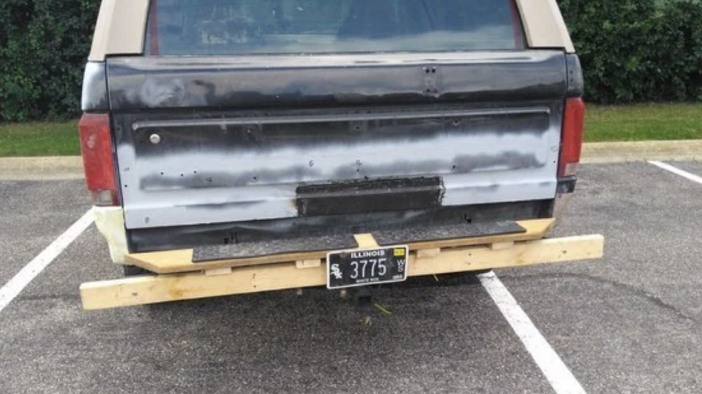 The image shows the back of a vehicle with various paint patches and a partially obscured Illinois license plate. The rear bumper has been replaced with a wooden plank spanning the width of the vehicle. The vehicle is parked in a parking lot.