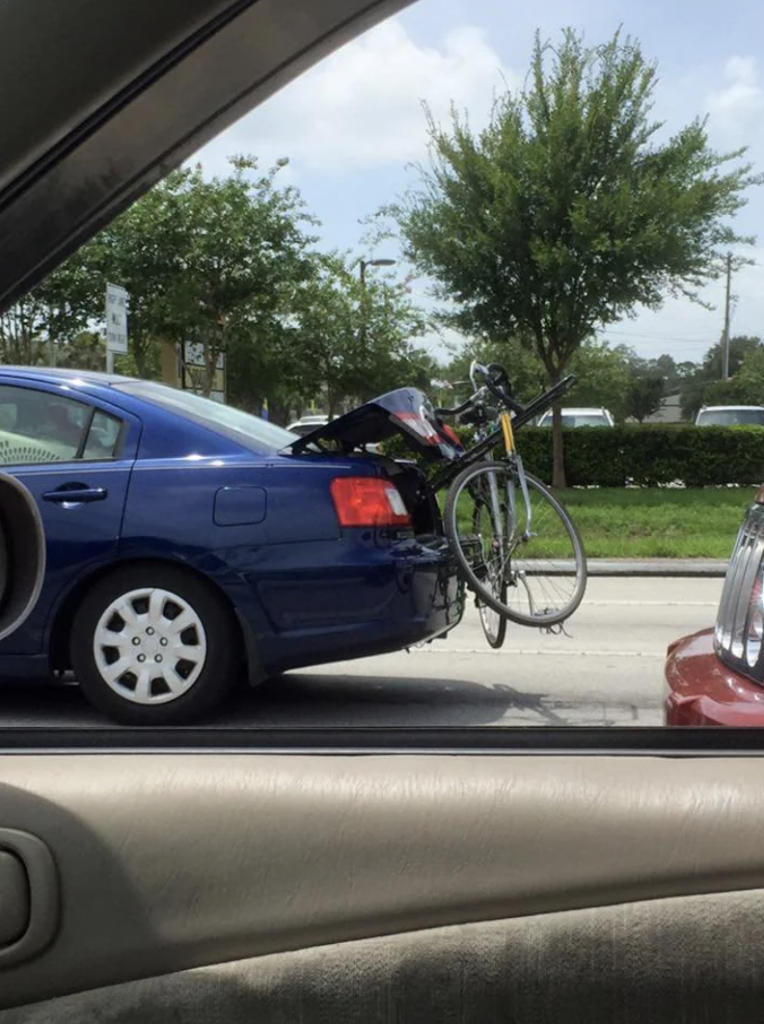 A blue sedan is seen on the road with a bicycle awkwardly attached to its partially open trunk. The bicycle's front wheel is hanging out, and the trunk is slightly open, secured with a bungee cord. Trees and other cars are visible in the background.
