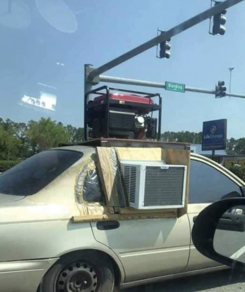 A beige sedan has an unconventional setup, with a window air conditioner unit installed on the rear side window. A red generator is secured to the car's roof, likely to power the AC. The vehicle is at a traffic light intersection on a clear day.