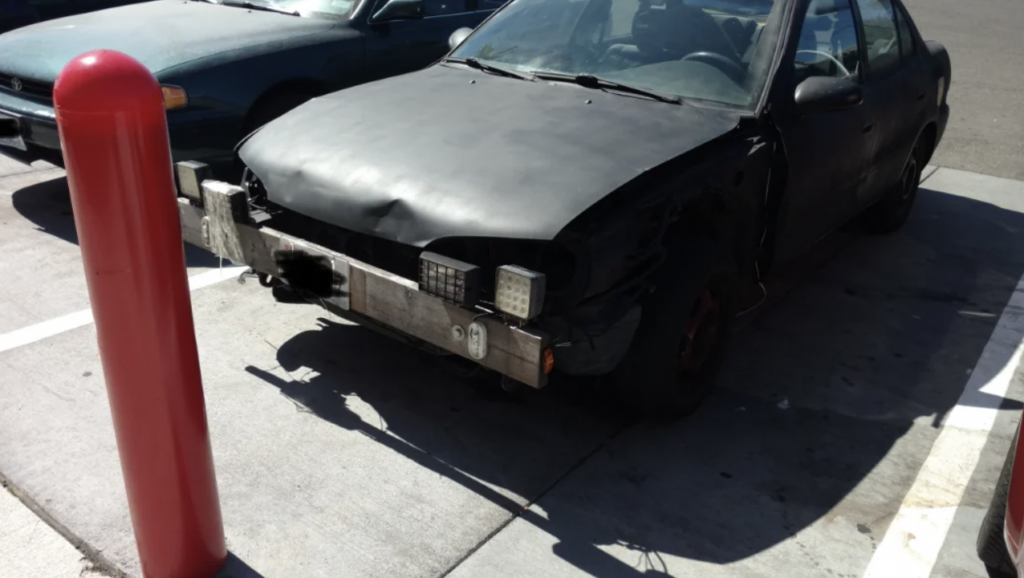 A heavily damaged car with a crumpled hood and missing front bumper is parked next to a red bollard on a sunny day. The car's headlights and front grill appear to be missing or broken, and the vehicle is in a state of disrepair.
