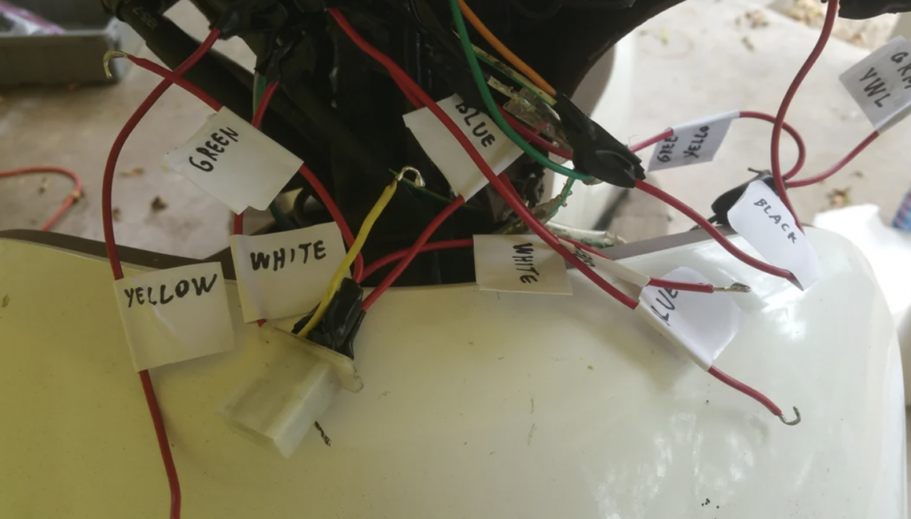 Close-up of numerous electrical wires bundled together, each tagged with a handwritten white label indicating color: green, white, blue, yellow, and black. The wires are connected to a white plug, possibly part of a larger machine or vehicle.