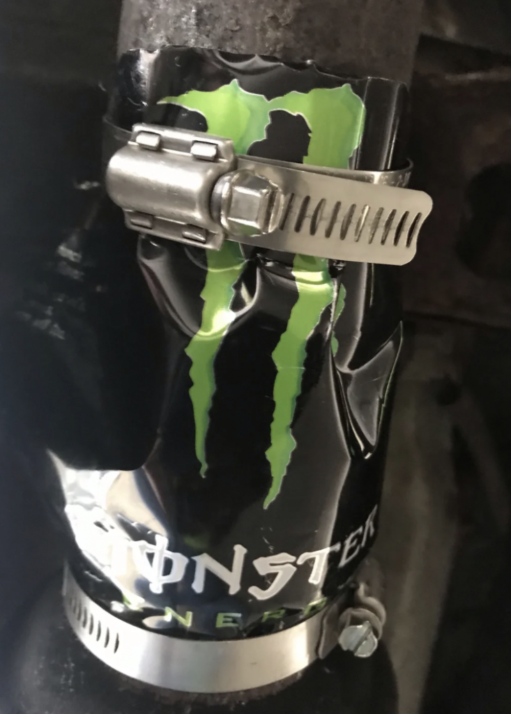 A close-up of a metal pipe secured with a hose clamp made from a cutout of a Monster Energy drink can. The can piece is wrapped around the pipe, and the green Monster logo is visible.