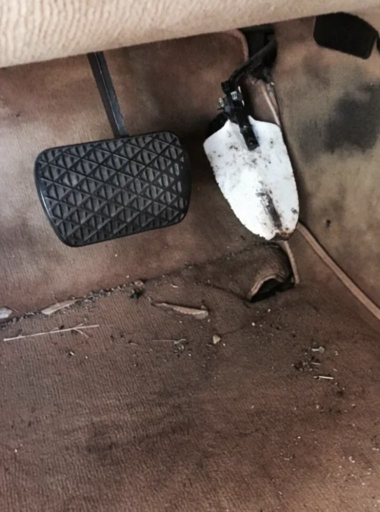 A worn car interior featuring a brake pedal and an accelerator pedal, both mounted on a dirty carpeted floor. The rubber brake pedal cover is intact, but the accelerator pedal is partially covered with a combination of white and black marks. The carpet shows signs of wear and debris.