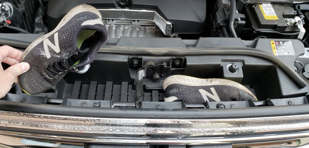 A person holds a pair of black New Balance running shoes in the engine compartment of a car. The car hood is open, revealing various engine components and the battery. The shoes are positioned as if they were placed on the engine components directly.