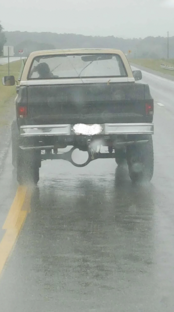 A black pickup truck with a beige top driving on a wet road in rainy weather. The license plate is blurred out. The truck appears to be lifted, and water spray is visible behind the tires. The road has double yellow lines and a safety barrier on the right.