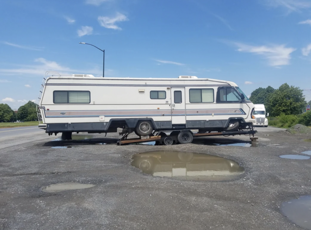 An RV is partially supported by a trailer on an uneven gravel surface. The rear wheels of the RV are on the ground, while the front is elevated. There are large puddles in the gravel. A white truck is parked off to the side in the background.