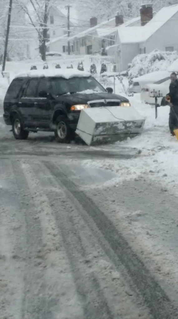 An SUV with a broken snowplow attachment sits in the middle of a snowy road. Snow-covered houses are visible in the background, and a person stands on the right side of the image, shoveling snow near a mailbox. Snow blankets the entire scene, creating a wintry landscape.