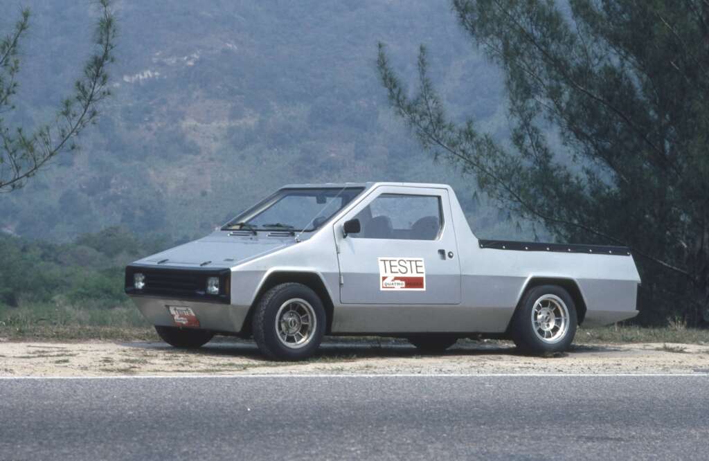 A silver, boxy, two-door vehicle labeled "TESTE" is parked along a road with trees and hills in the background. The car has a unique, angular design and a pickup-style bed. The scene appears to be captured in a natural, rural setting.