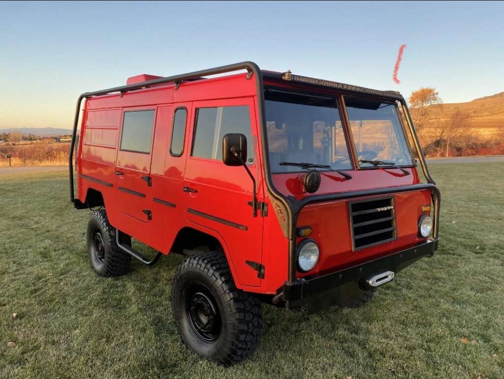 A red, rugged, off-road vehicle with large tires is parked on a grassy field. The vehicle has a boxy design, multiple windows, and black accents, including a roof rack and front bumper. The background features a distant fence, trees, and a clear sky at sunset.