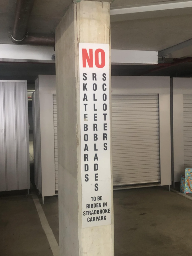 A vertical sign attached to a concrete pillar in a parking area reads, "NO SKATEBOARDS ROLLERBLADES SCOOTERS TO BE RIDDEN IN STRADBROKE CARPARK" in bold black and red text. The background shows metal storage units.