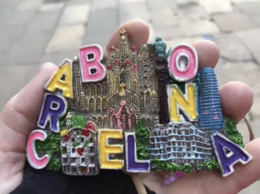 A hand holds a colorful refrigerator magnet spelling "Barcelona" with iconic landmarks like the Sagrada Familia, Park Güell, and the Torre Agbar depicted among the letters. The background is a blurred pavement.