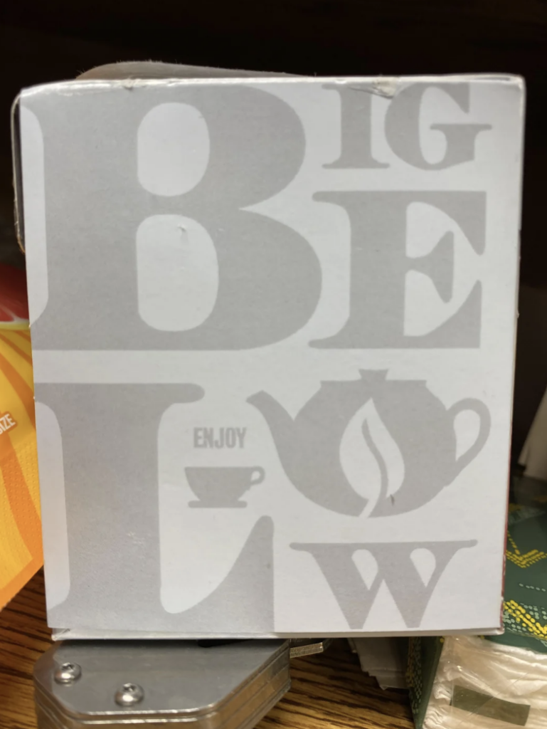 A close-up of a white box with large grey letters "BIGELOW" and images of a teapot, tea leaf, and teacup. The word "ENJOY" is between the teapot and teacup. Part of an orange box is visible on the left, and a green box can be seen on the right.