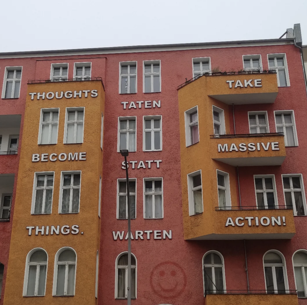 A colorful building with English and German text on its facade. Red and orange walls display the message: "THOUGHTS BECOME THINGS. TATEN STATT WARTEN. TAKE MASSIVE ACTION!" White-framed windows are spread across the building.