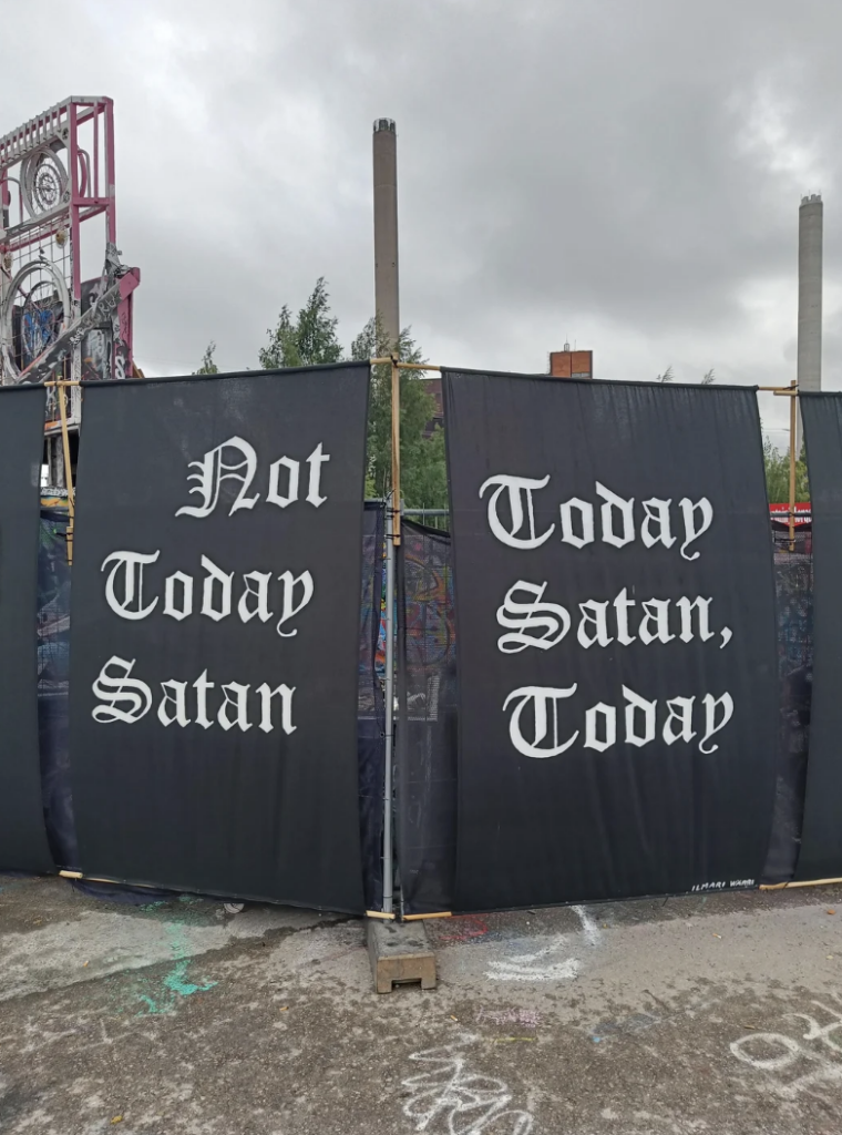 A gray, cloudy sky looms over a construction site with metal fences bearing black banners. The left banner reads "Not Today, Satan" in white gothic font, and the right banner says "Today Satan, Today" in the same font. The ground is covered in graffiti.